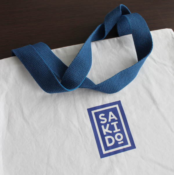 Gros plan d'un Tote bag MEIYO en toile de judogi blanche avec un logo Sakido bleu et des anses bleues.