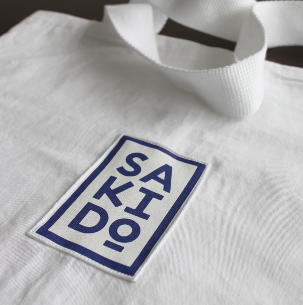 Tote bag MEIYO en toile de judogi blanche avec un logo Sakido bleu et des anses blanches.