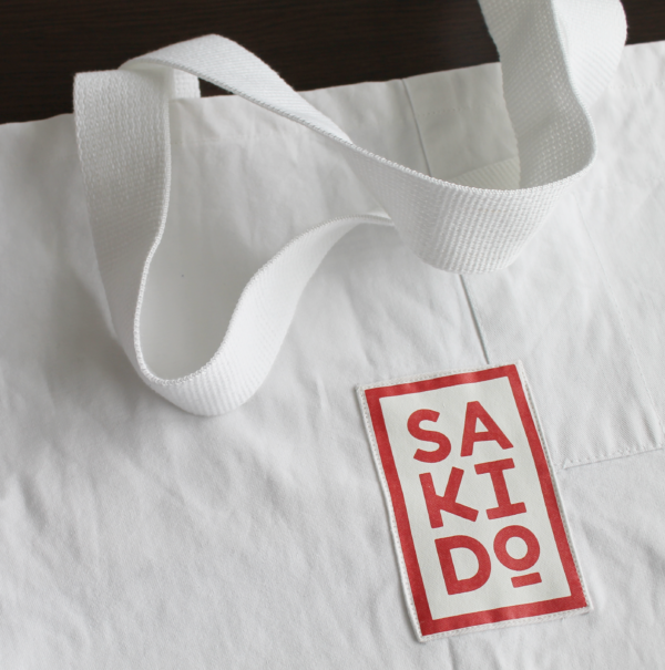 Gros plan d'un Tote bag MEIYO en toile de judogi blanche avec un logo Sakido rouge et des anses blanches.