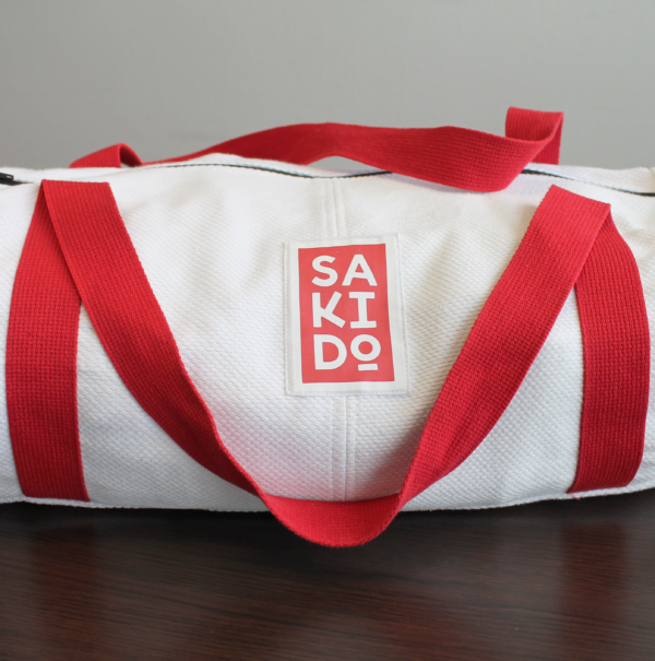 Un sac Polochon YUJO en toile de judogi avec une bandoulière rouge vif et un logo Sakido rouge, posé sur une table en bois.