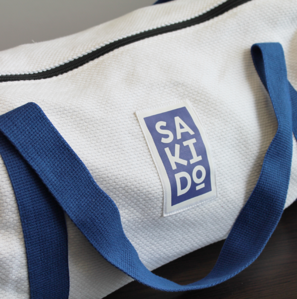 Un sac Polochon YUJO en toile de judogi avec une bandoulière bleu et un logo Sakido bleu, posé sur une table en bois.