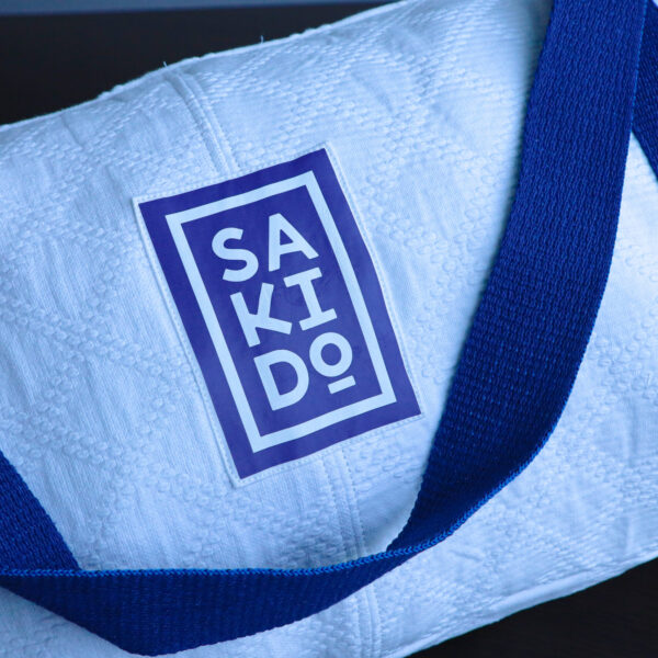 Gros plan d'un Sac Polochon YUJO, un sac en toile de judogi blanche avec une bandoulière bleue, comportant une étiquette logo Sakido bleu.